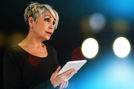 A woman with short hair talking and holding a tablet with lights in the background.
