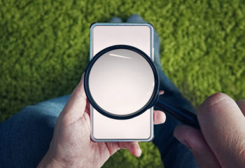 Man holding blank smartphone with magnifying glass above screen on grassy background.