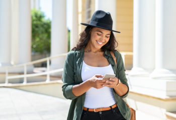 Happy woman wearing a hat using smartphone while traveling outside.