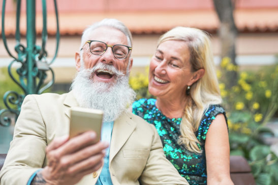 Happy senior couple watching on mobile smart phone and laughing together - Mature fashion people having fun with new technology cellphones outdoor - Social, pensioners, tech, elderly lifestyle concept