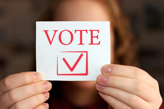 Image shows: Hands holding envelope with “VOTE” printed on it.
