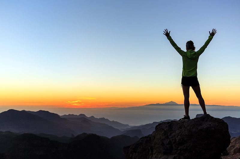 Image shows person with hands outstretched on mountain top looking at sunrise