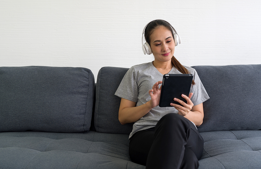 Woman, with tablet and white headphones, relaxes on dark sofa wile listening to audio magazines.